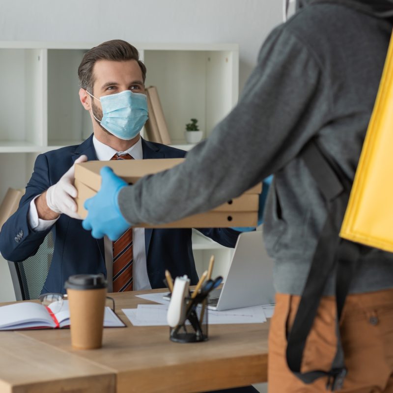 partial view of food delivery man giving pizza boxes to businessman in medical mask in office